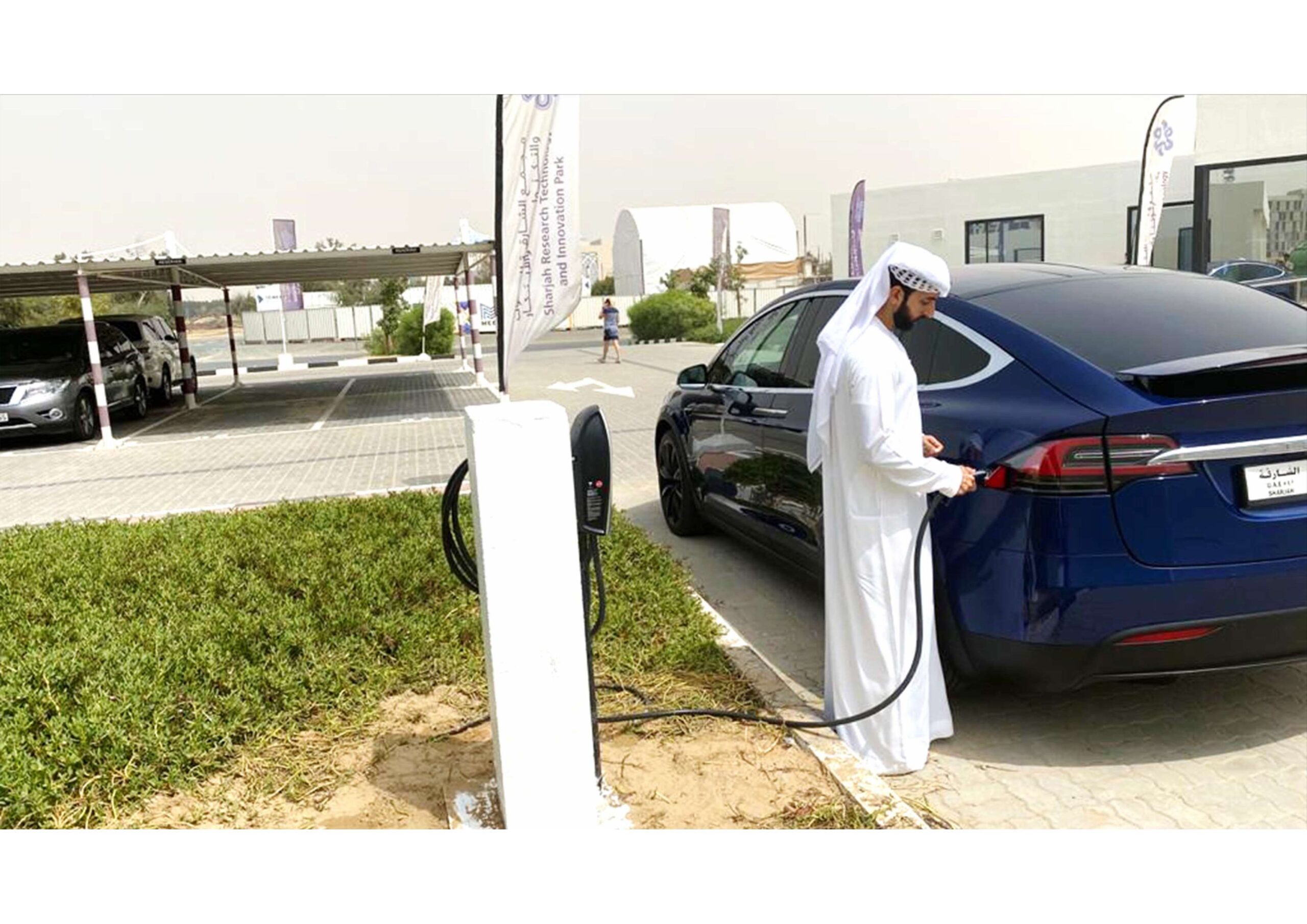 A man in traditional Middle Eastern attire charges a blue electric car at a charging station in a parking lot.