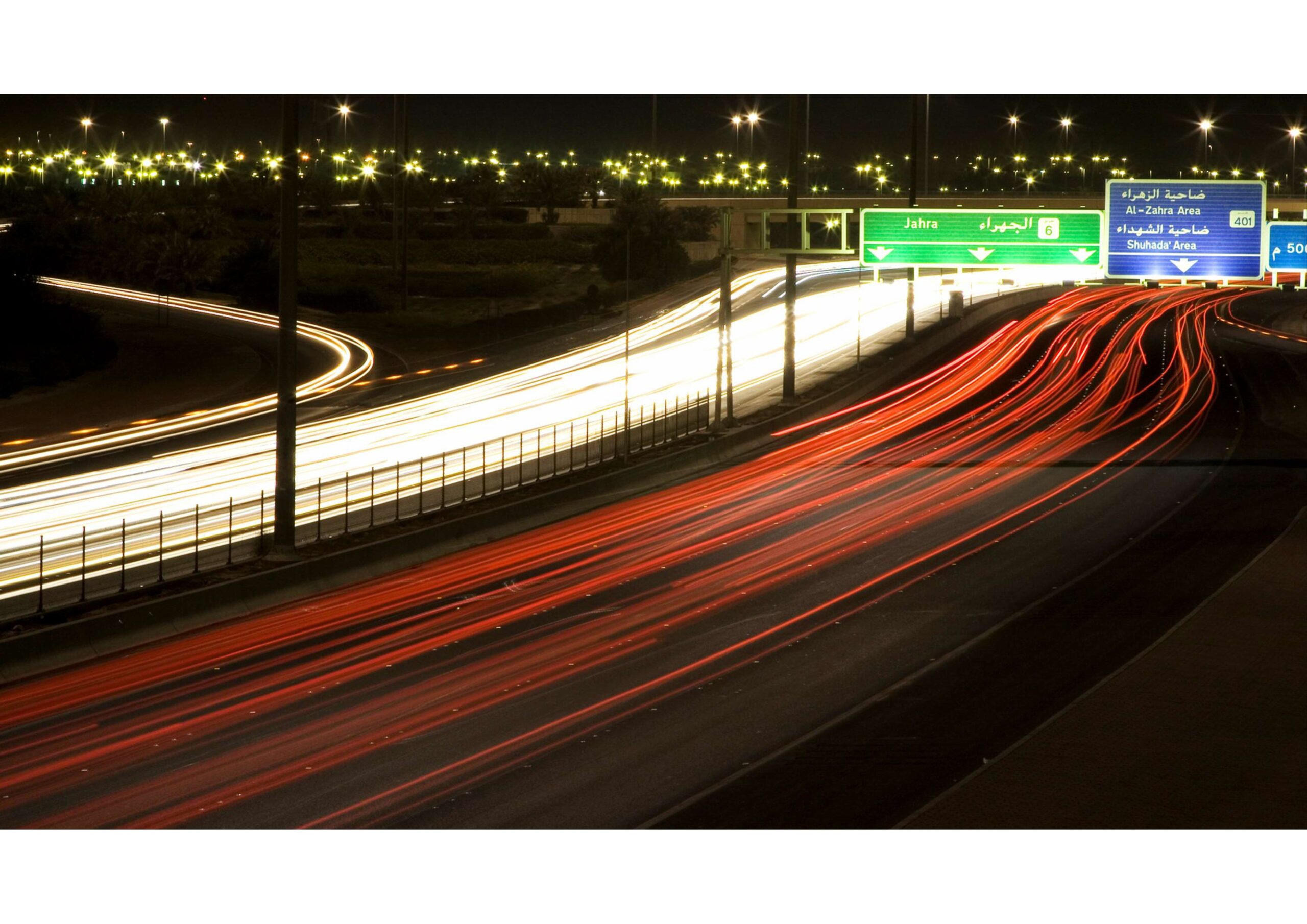 Long exposure photo of a busy highway at night, showing red and white light trails from vehicles, with green road signs in Arabic and English overhead.