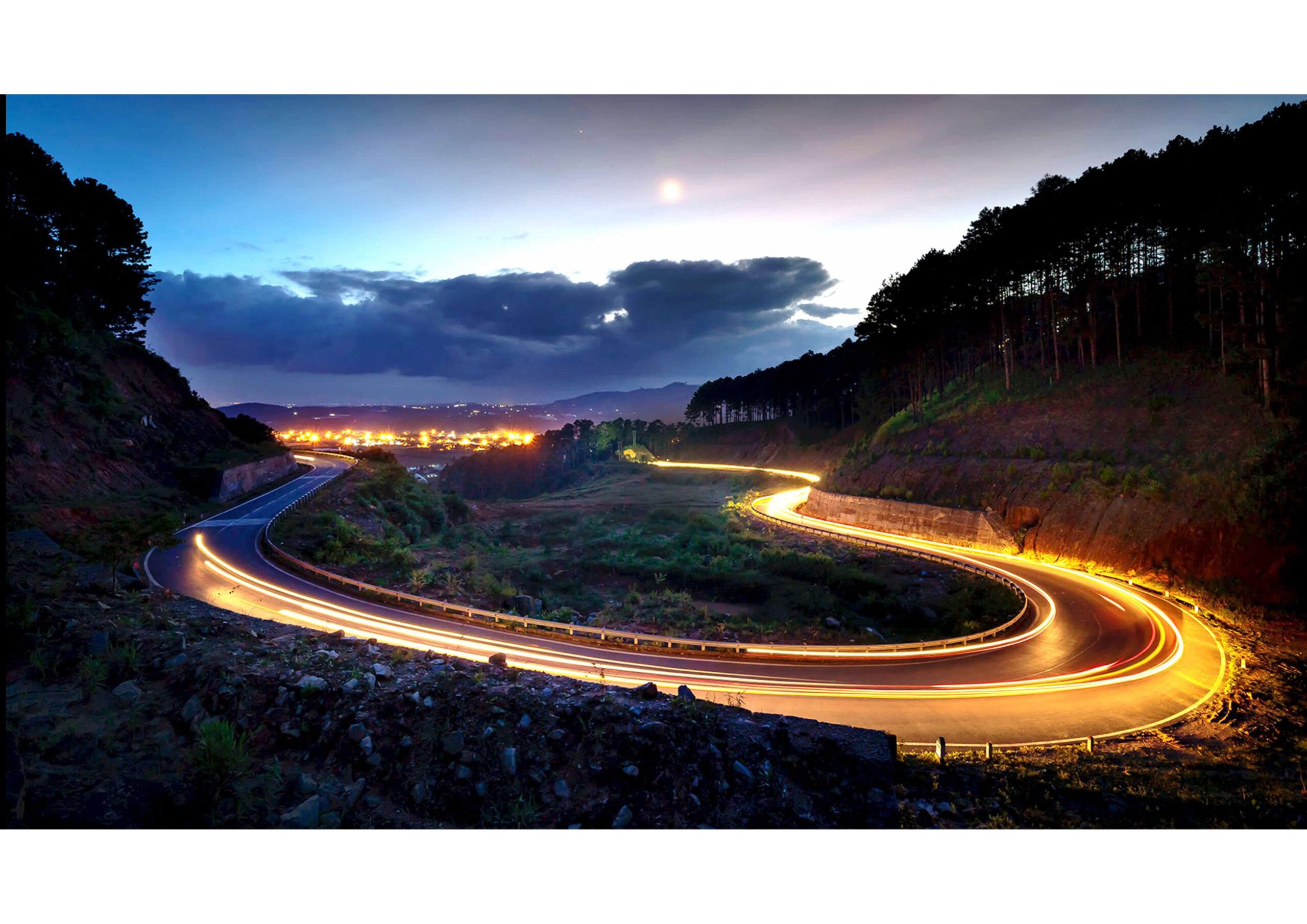 Curved mountain road at dusk with car light trails, city lights in the distance, silhouetted trees, and a cloudy sky with the moon visible.