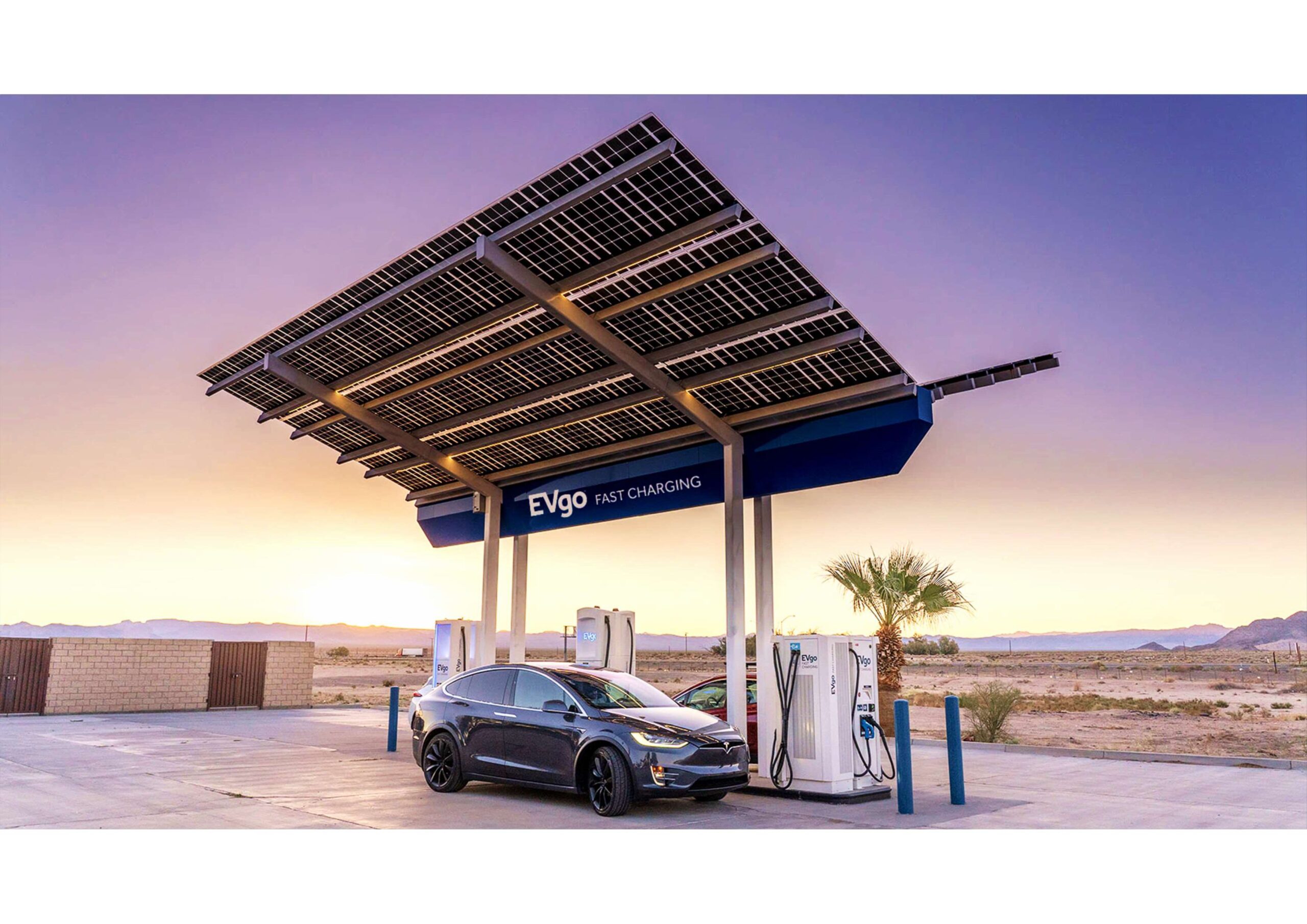 An electric vehicle is parked at an EVgo fast charging station beneath a solar panel canopy in a desert landscape at sunset.