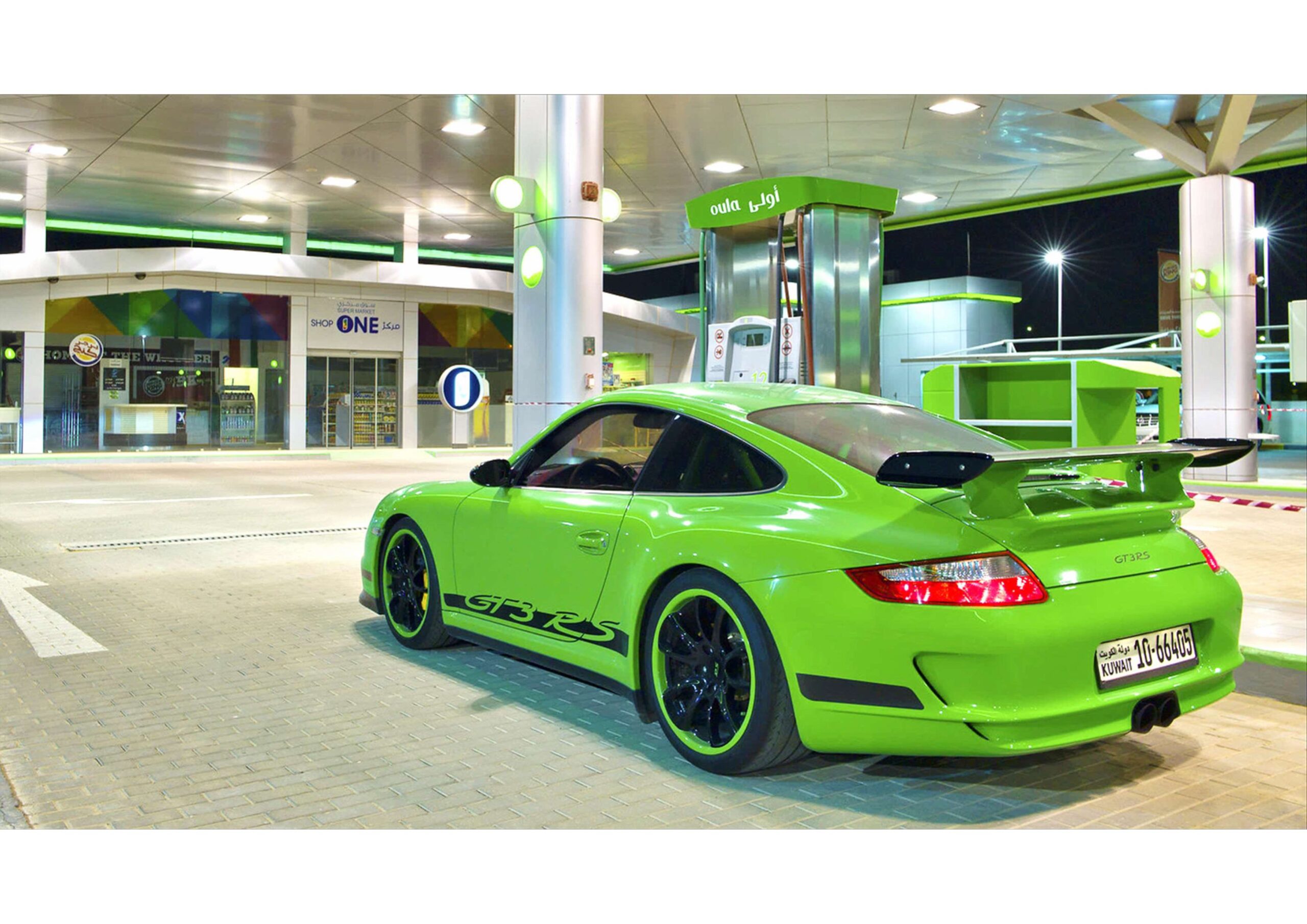 A bright green Porsche 911 GT3 RS is parked at a modern gas station at night, with fuel pumps and a well-lit building in the background.