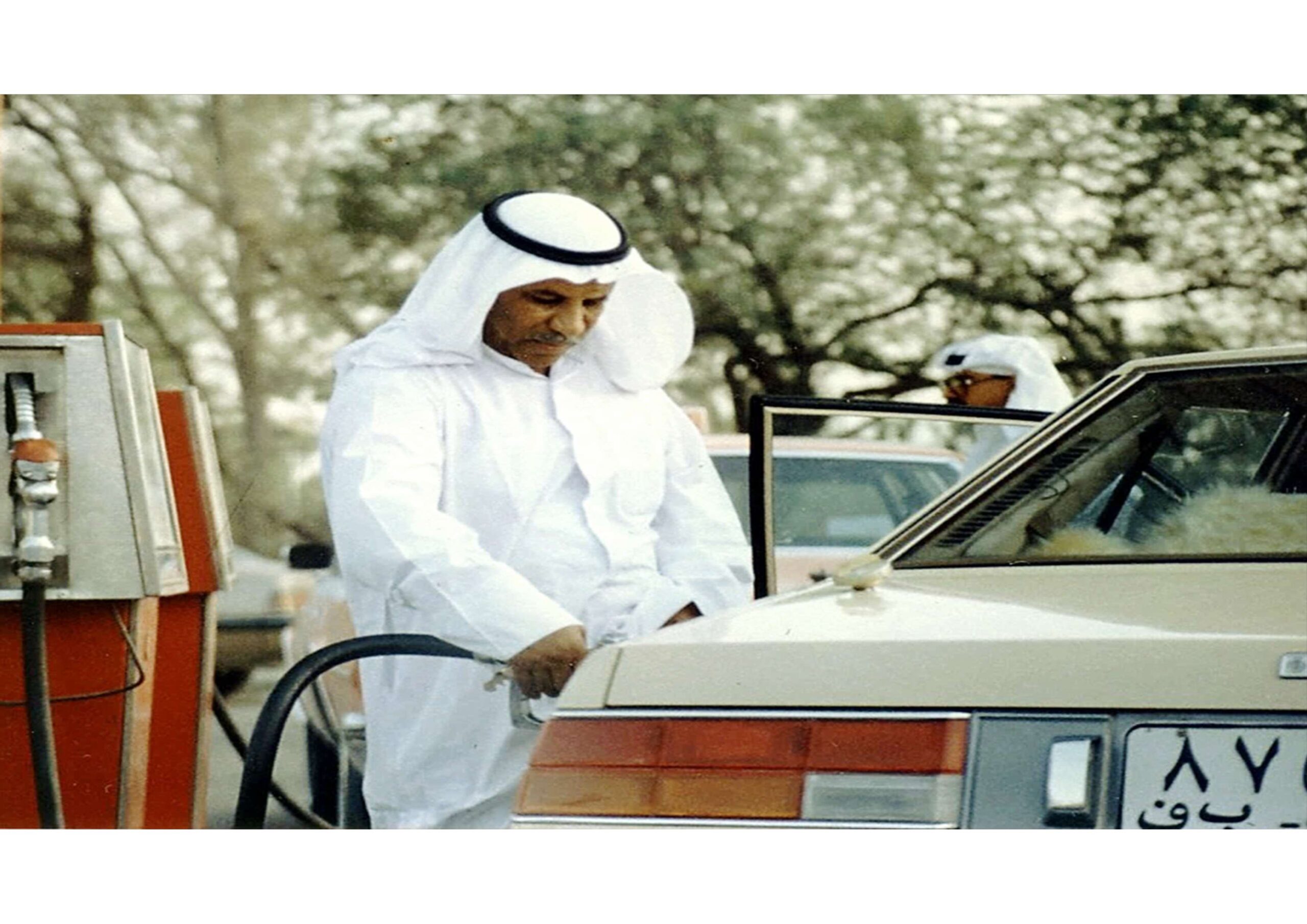 A man in traditional Middle Eastern attire pumps gasoline into a beige car at an outdoor gas station.