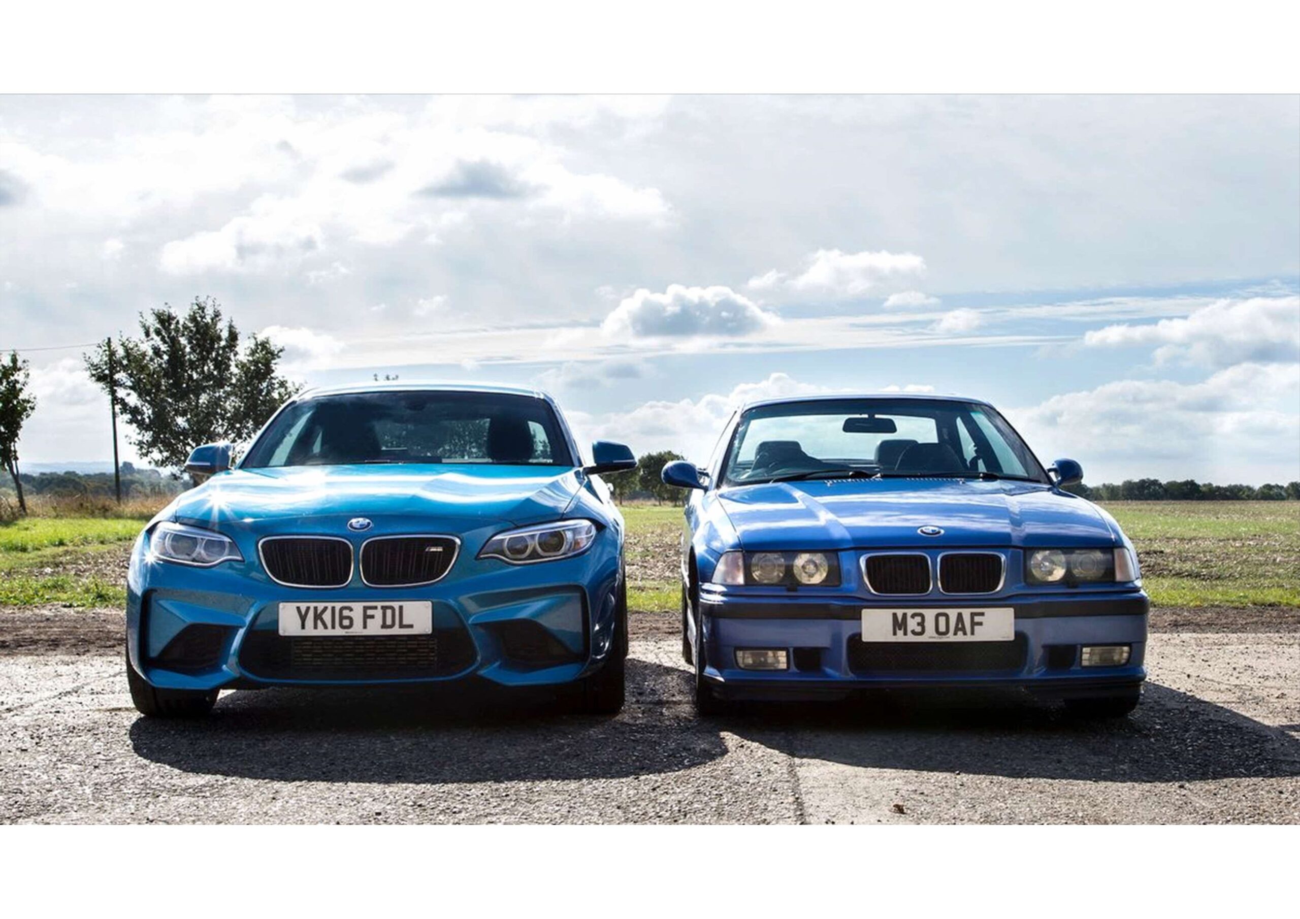 Two blue BMW cars are parked side by side on a paved area outdoors, with a field and cloudy sky in the background.