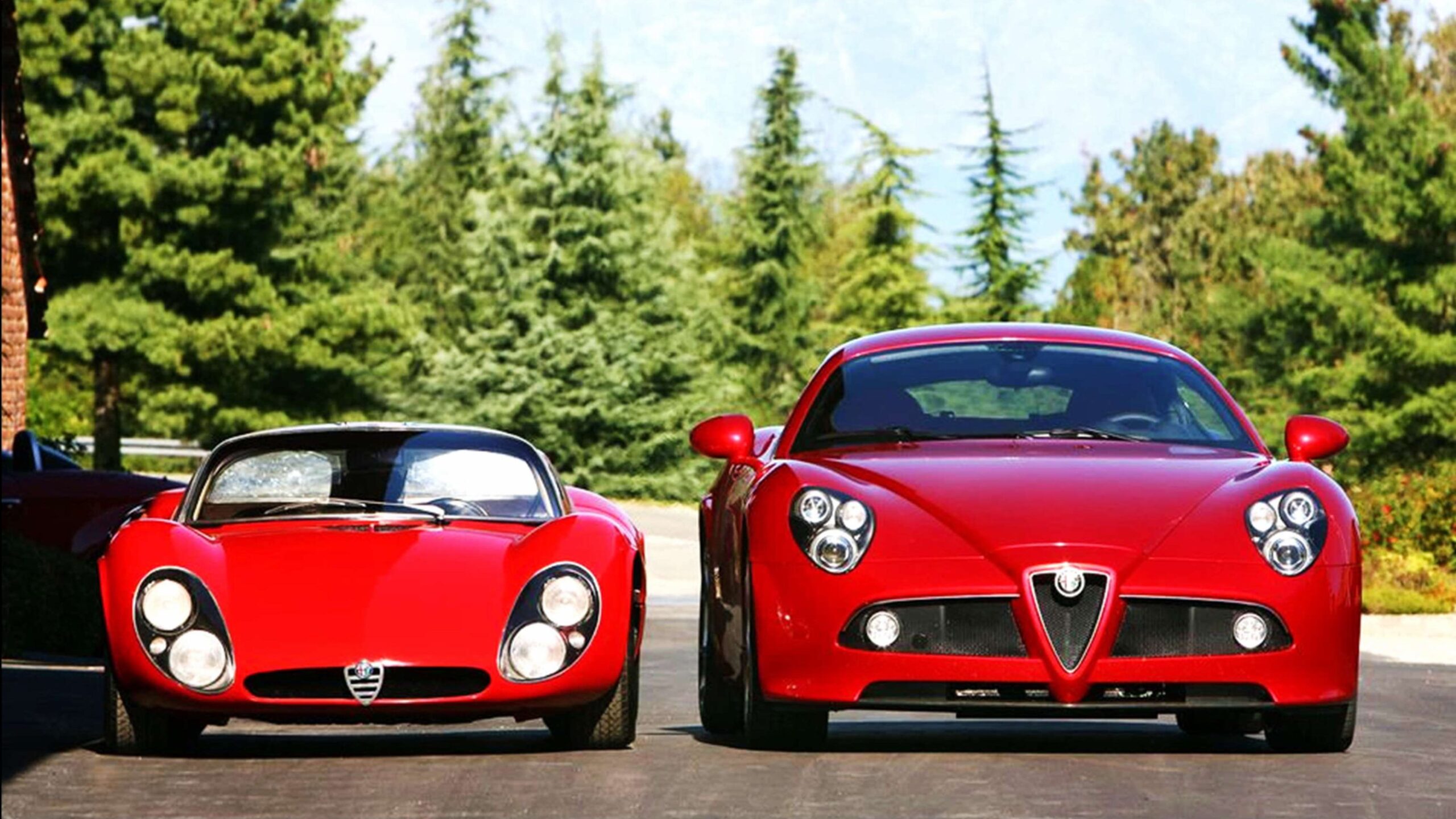 Two red Alfa Romeo sports cars are parked side by side on a road with trees and mountains in the background.