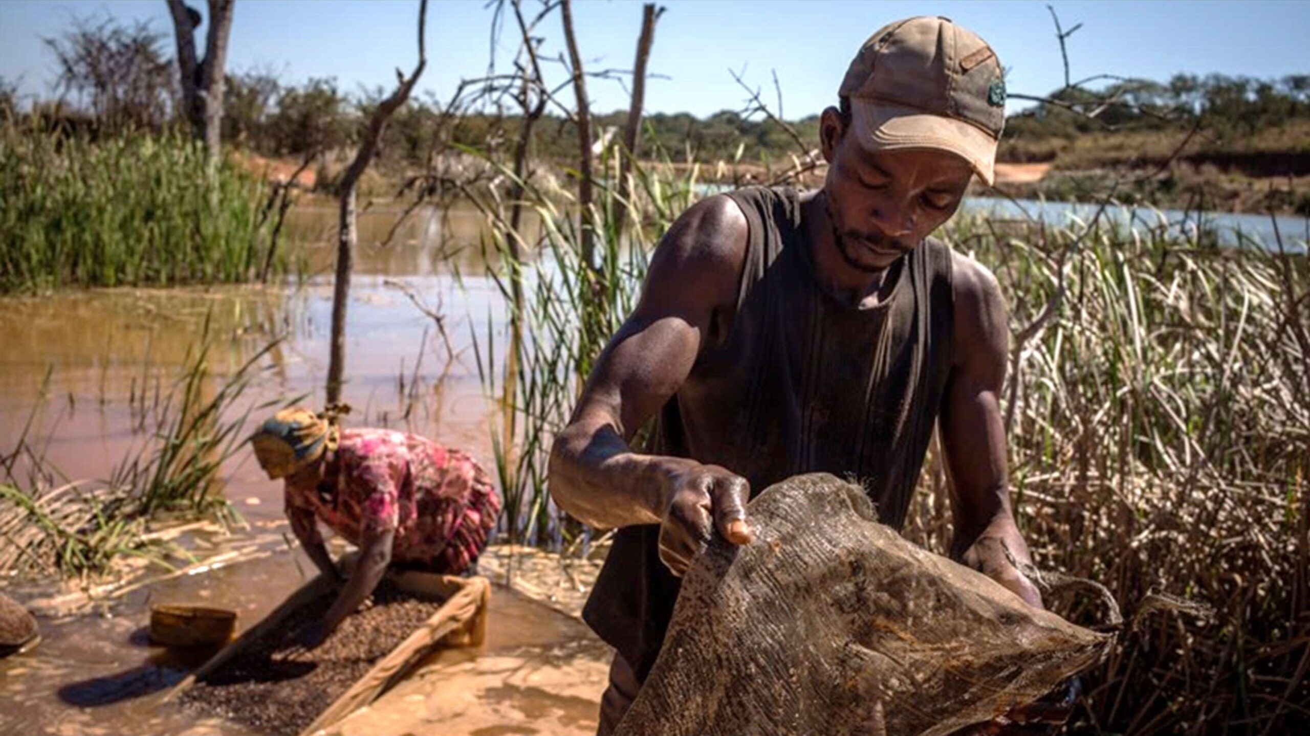 Aerial view of rectangular evaporation ponds in various shades of green, blue, and yellow, separated by dirt roads in a dry, industrial landscape.