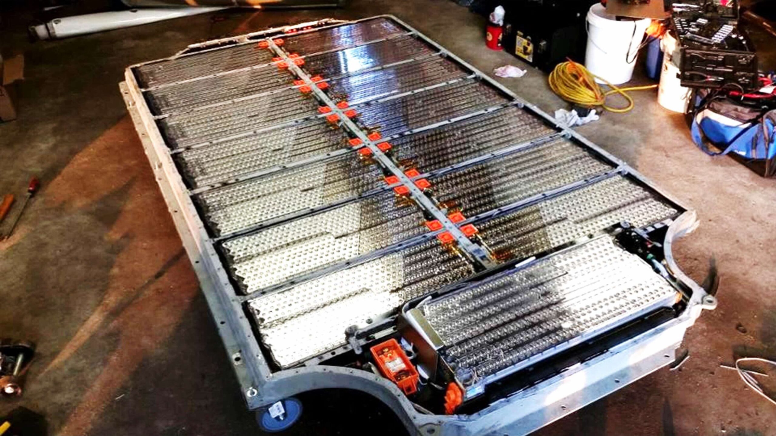 A large electric vehicle battery pack is disassembled and laid out on a garage floor, showing its internal modules and wiring.