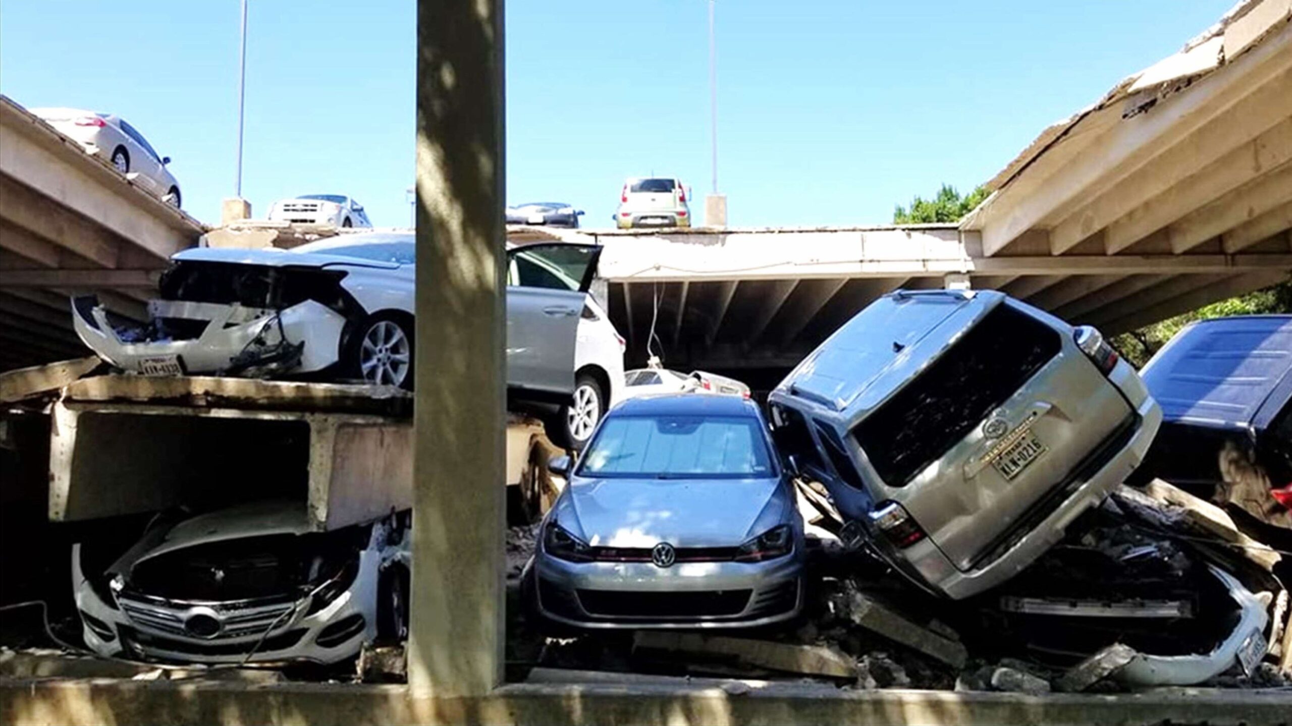 Several cars are crushed and stacked on top of each other beneath a partially collapsed overpass, with more vehicles on the damaged upper roadway.