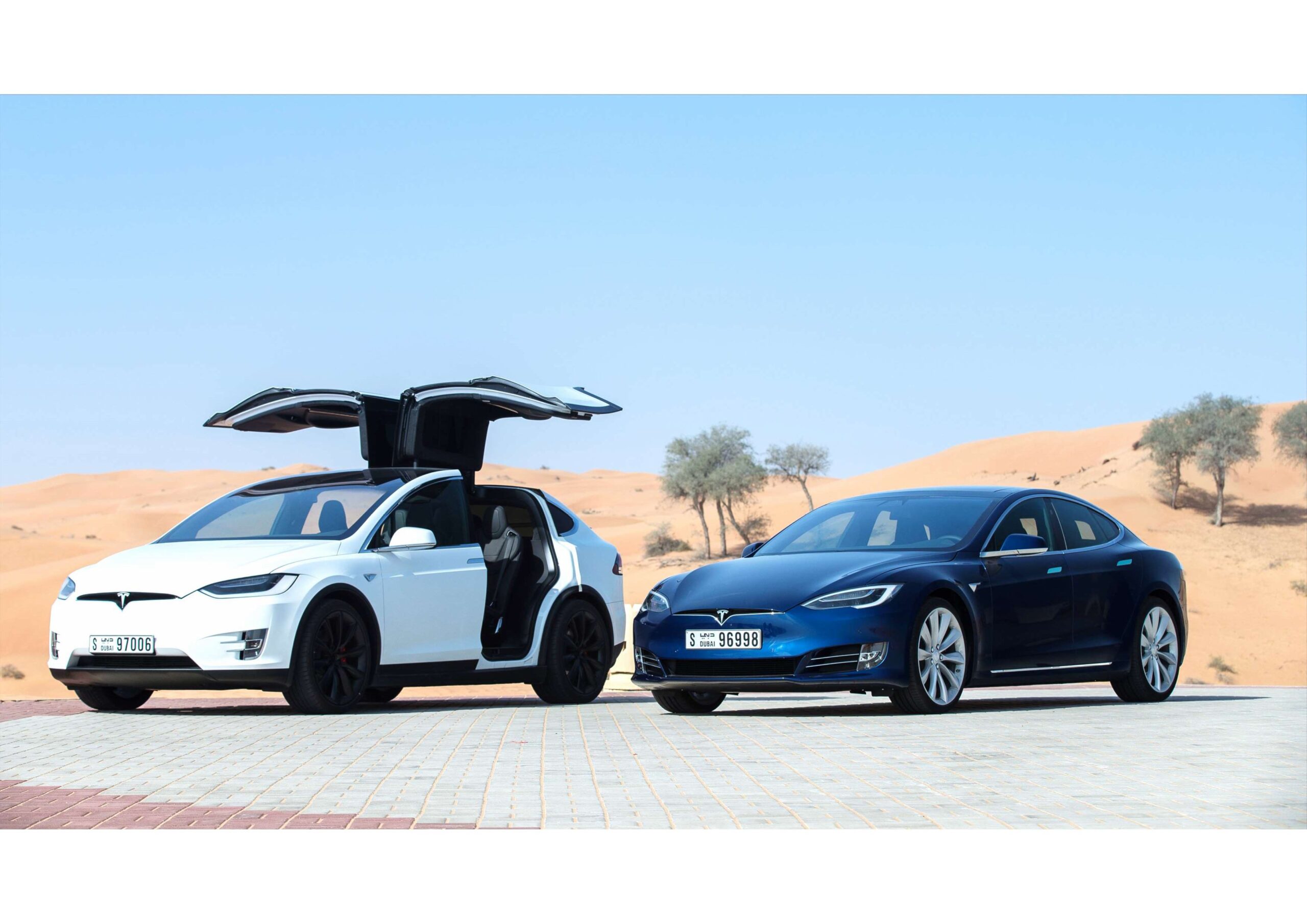 A white Tesla SUV with its falcon wing doors open and a blue Tesla sedan are parked side by side on a paved surface in a desert landscape.