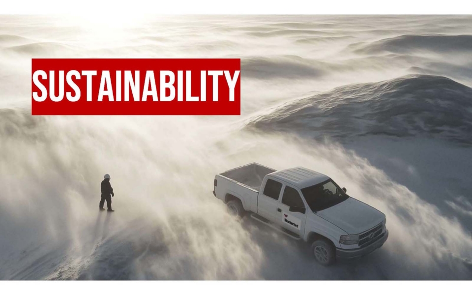 A person stands next to a white pickup truck on a snowy, windswept landscape with the word “SUSTAINABILITY” in bold red and white text above.