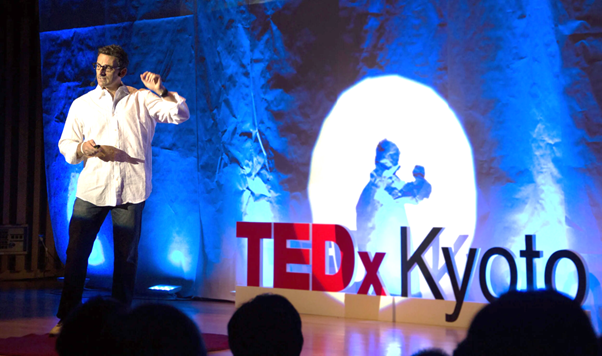 A speaker in a white shirt presents on stage at TEDxKyoto, with blue lighting and a large TEDxKyoto sign in the foreground.